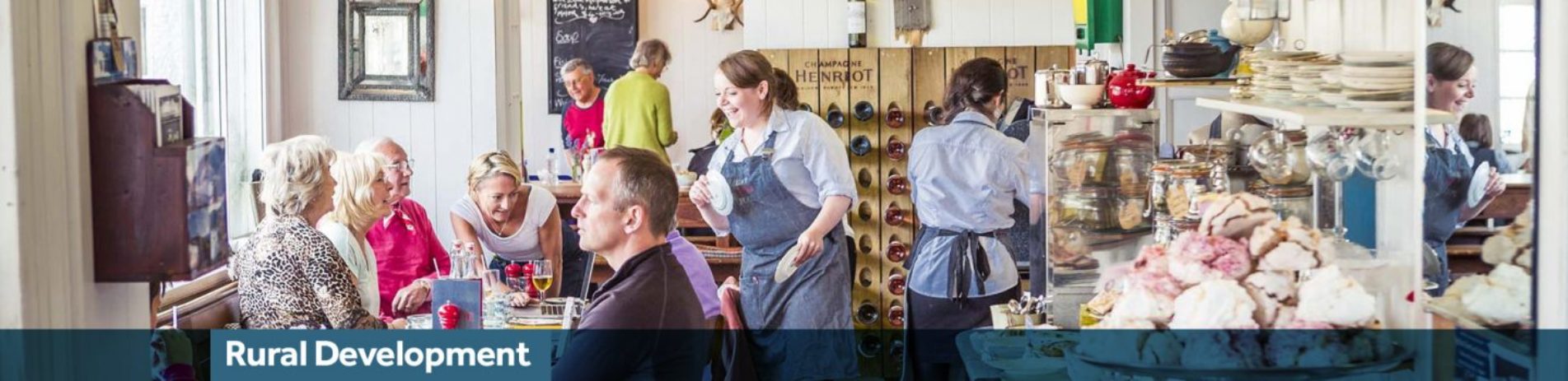 busy-tearoom-with-smiling-shop-assistant-chatting-to-seated-group-colourful-cakes-are-visible-on-right-and-blue-banner-underneath-reading-rural-development