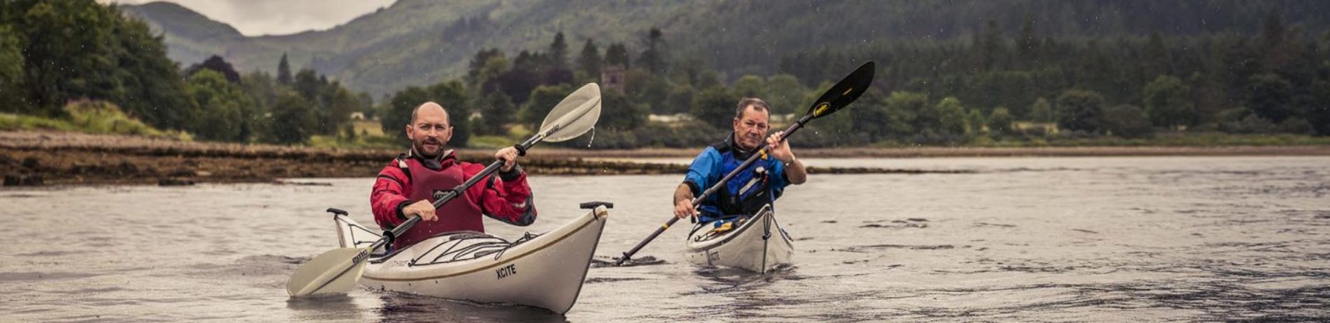 two-men-one-in-red-the-other-in-blue-peddling-in-separate-kayaks-on-loch-long-with-ardentinny-in-the-distance