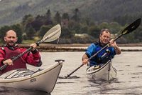 two-men-one-in-red-the-other-in-blue-peddling-in-separate-kayaks-on-loch-long-with-ardentinny-in-the-distance