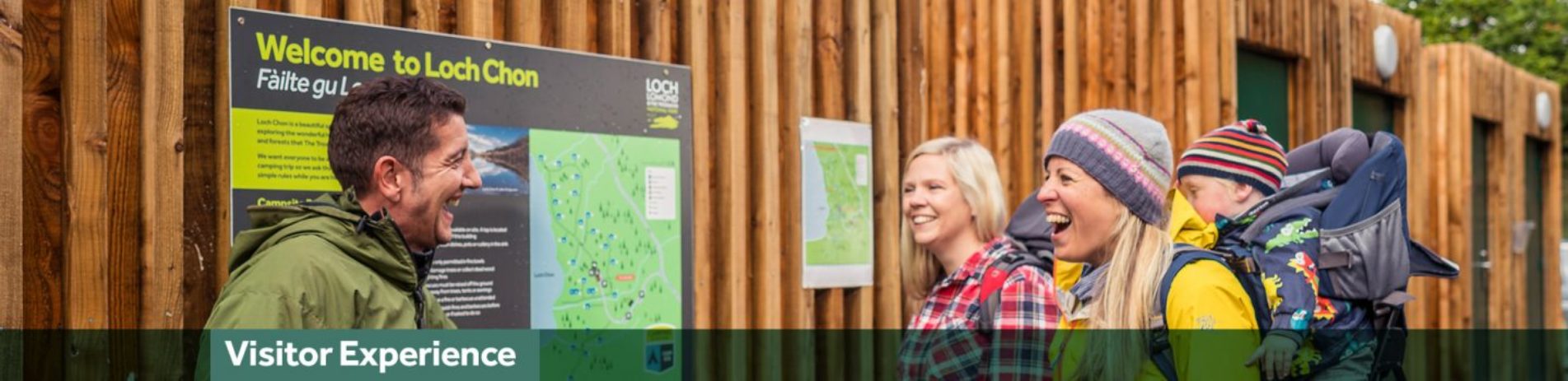 ranger-in-green-jacket-talking-enthusiastically-to-two-blonde-women-one-with-a-baby-on-her-back-at-the-loch-chon-campsite-with-green-banner-underneath-reading-visitor-experience