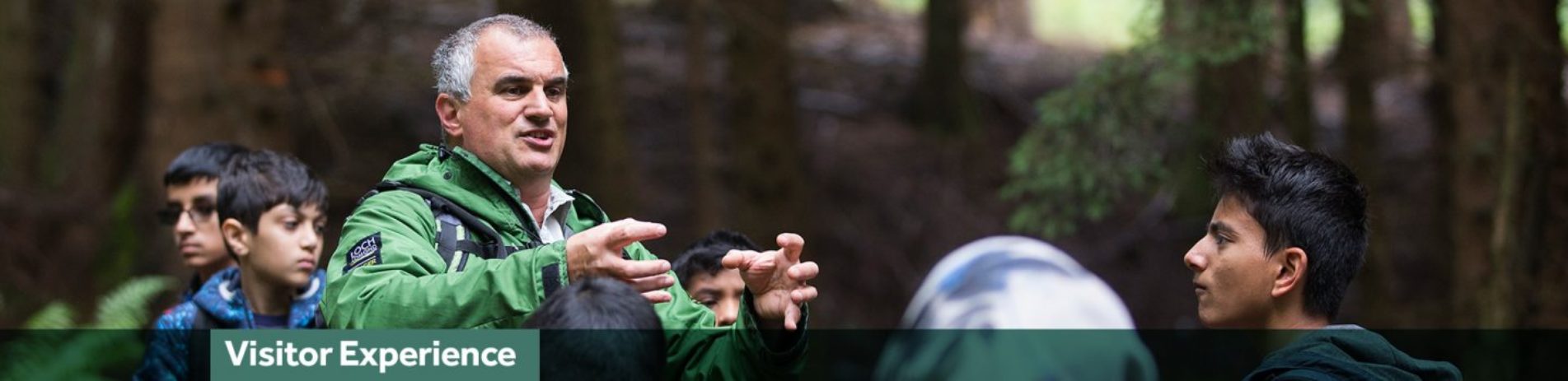 national-park-ranger-in-green-jacket-demonstrates-something-using-his-hands-in-front-of-school-students-in-forest-with-green-banner-underneath-reading-visitor-experience