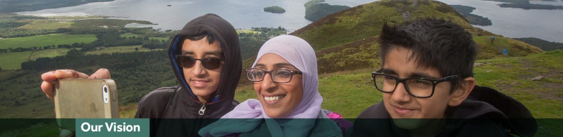 woman-wearing-white-hijab-and-glasses-and-with-two-young-boys-with-glasses-takes-selfie-loch-lomond-visible-behind-them-and-blue-banner-underneath-reads-our-vision