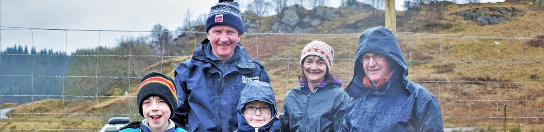 achray-farm-owners-posing-with-two-young-students-next-to-deer-fence-all-wearing-waterproofs-as-its-raining