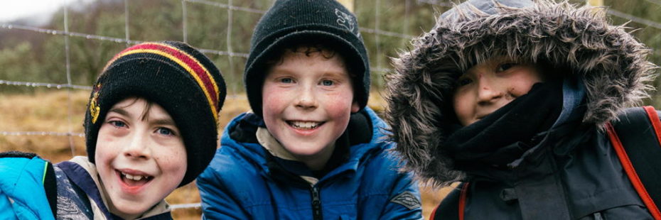 three-kids-in-thick-coats-and-hats-and-hoods-posing-next-to-deer-fence-at-loch-achray-farm