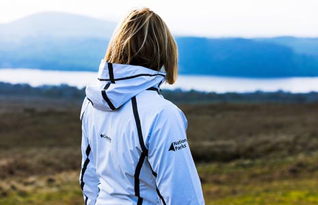 blonde-woman-in-white-columbia-jacket-imprinted-with-national-park-arrow-symbol-gazing-into-distance-to-loch