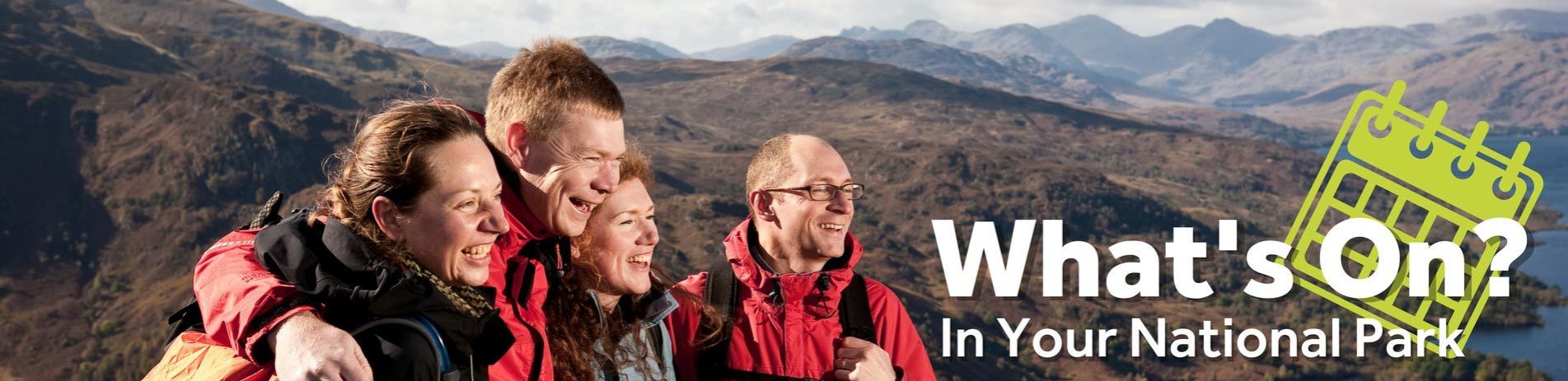 four-friends-two-male-two-female-holding-each-other-around-the-shoulder-and-laughing-on-summit-of-ben-aan-trossachs-mountains-and-loch-katrine-behind-them-text-in-corner-whats-on-in-your-national-park-with-green-calander-graphic