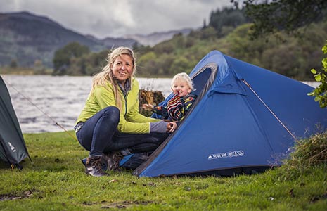 blonde-woman-stooped-next-to-blue-tent-entrance-holding-blond-baby-boy-loch-chon-behind