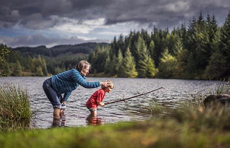 woman-with-child-ankles-in-water-loch-drunkie-three-lochs-forest-drive-coniferous-forests-visible-in-the-background