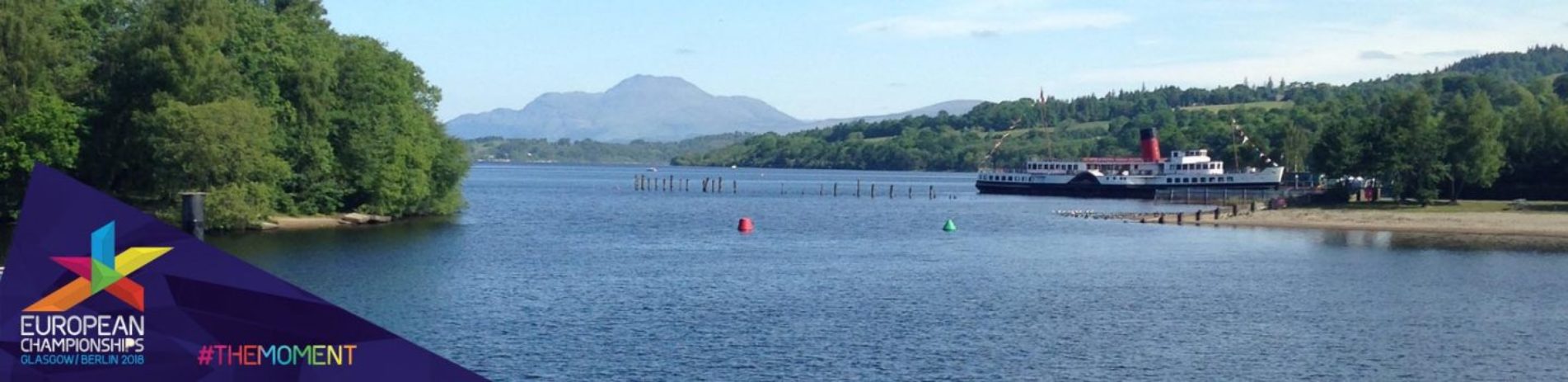 view-of-loch-lomond-wooded-shores-maid-of-the-loch-steamship-and-ben-lomond-in-the-distance-with-colourful-logo-of-european-championships-two-thousand-eighteen-in-bottom-left-corner