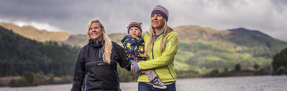 two-blonde-women-in-outdoor-gear-holding-baby-at-edge-of-loch-chon-with-hills-and-forests-behind-them