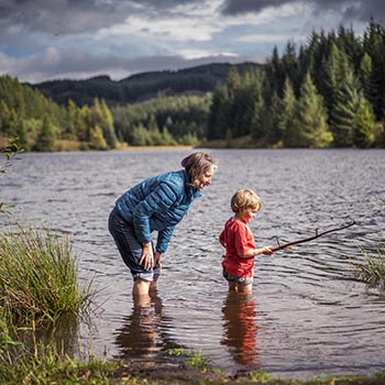 woman-with-child-ankles-in-water-loch-drunkie-three-lochs-forest-drive-coniferous-forests-visible-in-the-background