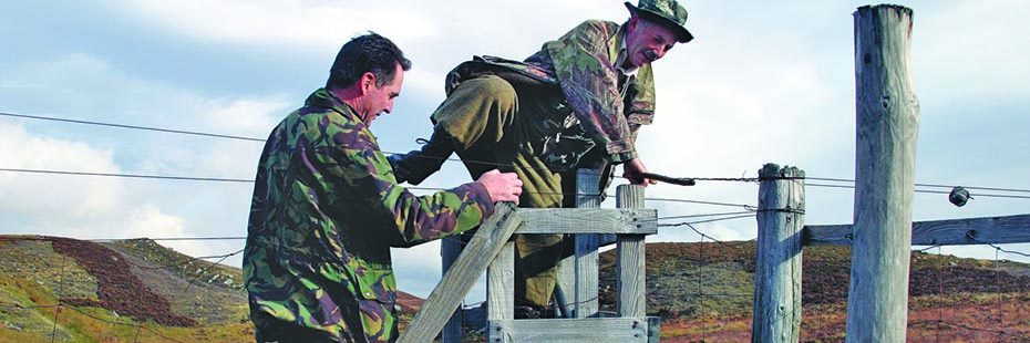 two-men-in-khaki-camouflage-clothes-climbing-deer-fence-moors-behind