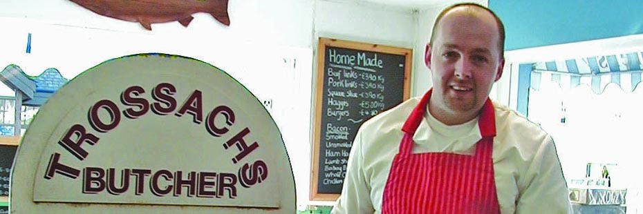 man-in-red-apron-in-shop-with-trossachs-butcher-sign-and-price-list-in-chalk-on-the-wall-next-to-him