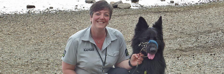 emma-white-volunteer-ranger-in-branded-grey-shirt-short-brown-hair-and-smiling-crouching-on-ground-next-to-large-black-dog-on-loch-beach