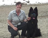 emma-white-volunteer-ranger-in-branded-grey-shirt-short-brown-hair-and-smiling-crouching-on-ground-next-to-large-black-dog-on-loch-beach