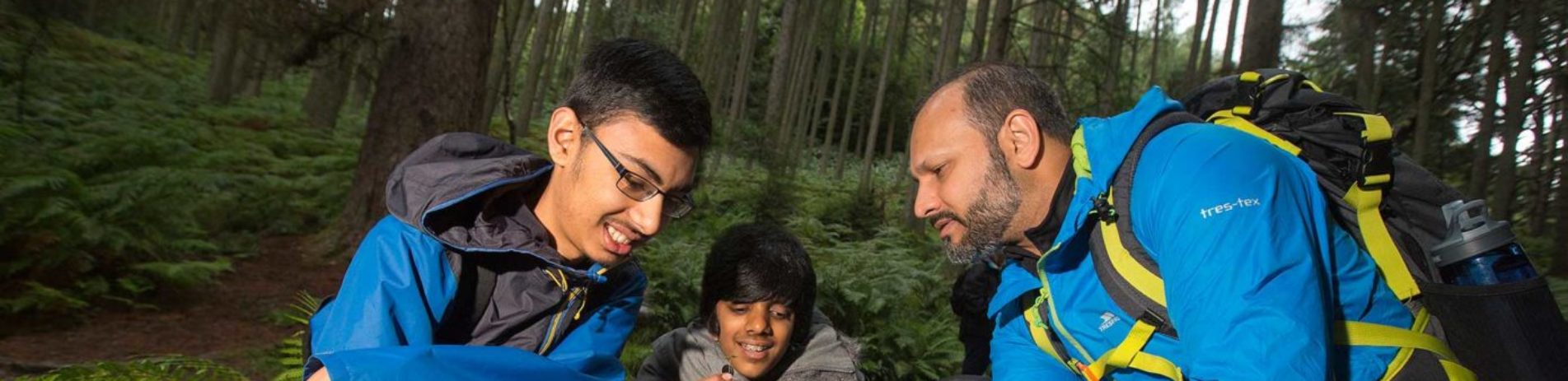 middle-aged-bearded-man-in-blue-outdoor-jacket-and-rucksack-crouched-on-ground-with-two-male-pupils-showing-them-a-plant-in-his-fingers-close-to-the-ground-they-are-in-the-forest