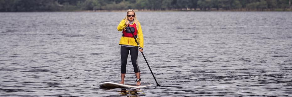 blonde-woman-in-yellow-jacket-and-wearing-sunglasses-paddleboarding-on-loch-lomond