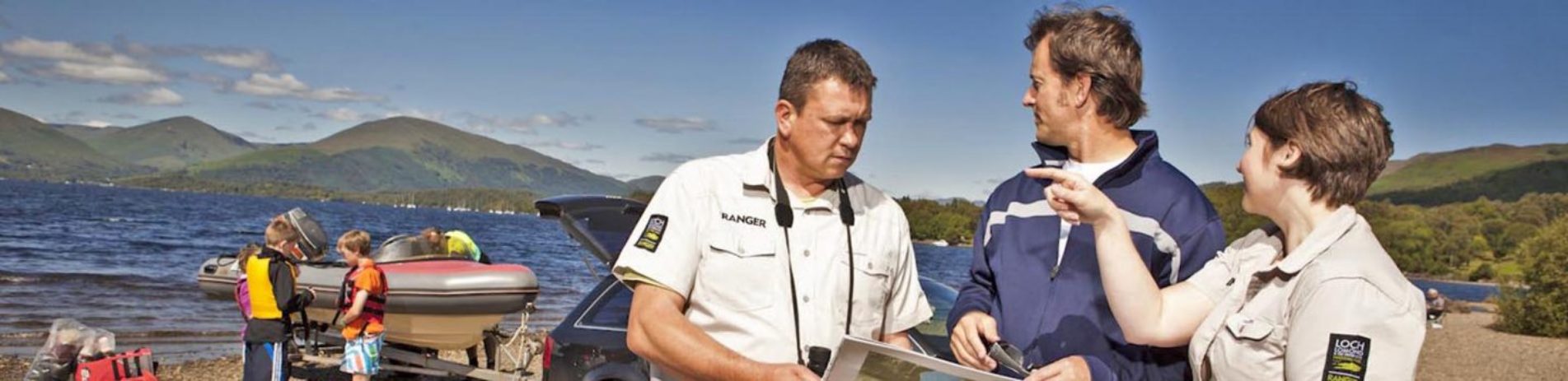 two-rangers-talking-to-member-of-the-public-and-advising-him-using-map-at-milarrochy-bay-on-summer-day-boat-launching-behind