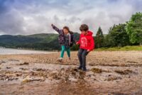girl-and-boy-in-colourful-clothes-holding-hands-jumping-in-stream-at-edge-of-loch-long-in-ardentinny