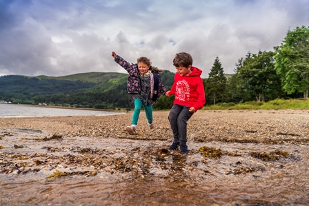 girl-and-boy-in-colourful-clothes-holding-hands-jumping-in-stream-at-edge-of-loch-long-in-ardentinny