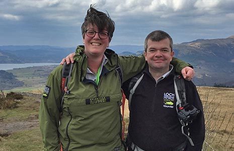two-volunteer-rangers-in-green-and-black-jackets-smiling-at-the-camera-with-windy-landscape-behind