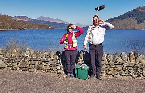 two-volunteer-rangers-one-female-and-one-male-with-bucket-and-brush-standing-erect-and-mock-saluting-militarily-at-the-camera-with-loch-and-hills-in-the-back-and-blue-skies