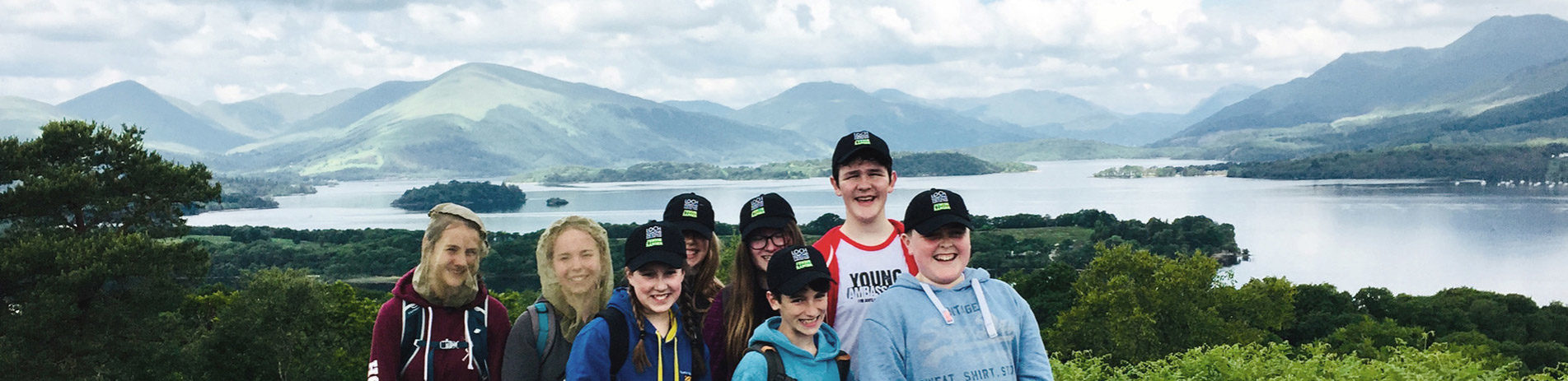young-volunteers-with-national-park-some-wearing-midge-nets-smiling-for-camera-at-summit-of-inchcailloch-with-stunning-views-behind-of-island-loch-lomond-and-surrounding-hills-including-ben-lomond