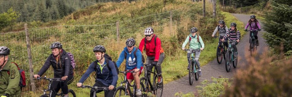 group-of-young-people-along-national-park-ranger-on-bikes-kitted-up-and-with-helmets-on-cycling-on-national-cycle-route-number-seven-in-glen-ogle-next-to-viaduct-surrounded-by-lush-greenery