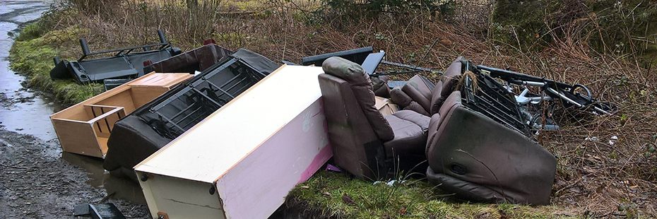furtniture-including-shelves-and-sofas-fly-tipped-at-edge-of-road-with-bracken-and-puddles-visible-around