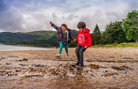 girl-and-boy-in-colourful-clothes-holding-hands-jumping-in-stream-at-edge-of-loch-long-in-ardentinny