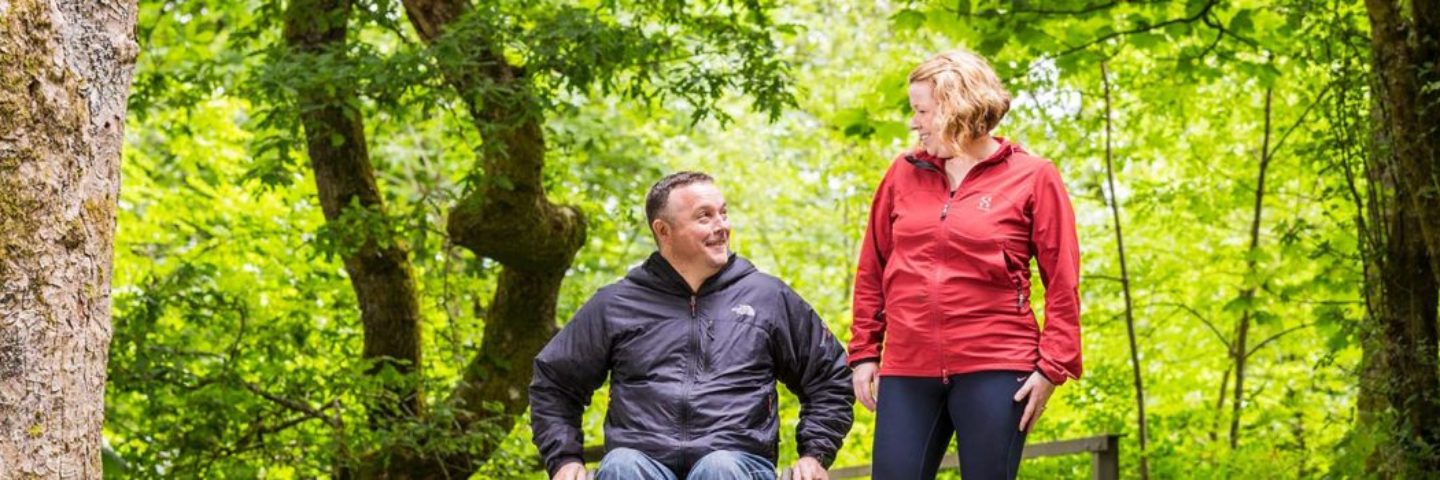 man-in-wheelchair-wearing-dark-grey-jacket-and-woman-in-jogging-clothes-including-red-top-on-path-in-balloch-castle-country-park-surrounded-by-lush-greenery