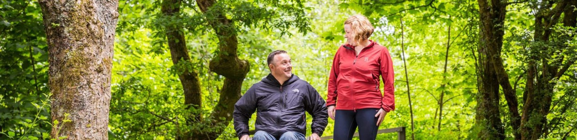 man-in-wheelchair-wearing-dark-grey-jacket-and-woman-in-jogging-clothes-including-red-top-on-path-in-balloch-castle-country-park-surrounded-by-lush-greenery