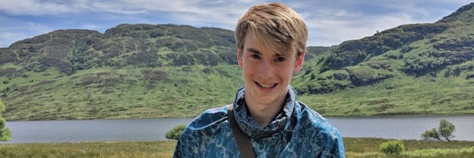struan-burch-volunteer-with-national-park-blond-hair-and-blue-shirt-against-landscape-of-loch-and-hills