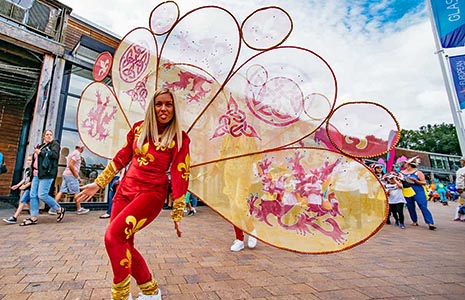 blonde-woman-dressed-in-red-and-yellow-with-fleur-de-lis-imprints-wearing-butterfly-like-wings-on-back-at-lomond-shores-during-balloch-festival-two-thousand-eighteen