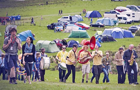 group-of-people-some-playing-instruments-walking-up-slope-with-dozens-of-tents-and-campervans-in-the-distance-mhor-festival