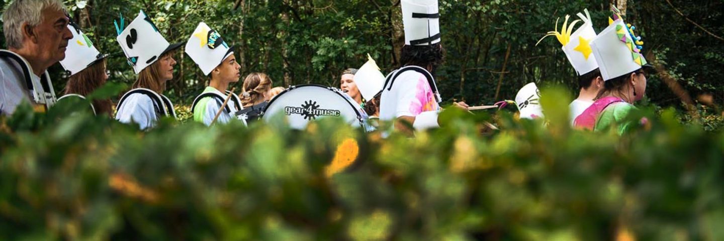 group-playing-drums-in-procession-and-wearing-tall-white-hats-beyond-green-hedges-at-balloch-festival-two-thousand-eighteen