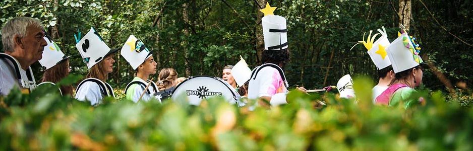 group-playing-drums-in-procession-and-wearing-tall-white-hats-beyond-green-hedges-at-balloch-festival-two-thousand-eighteen