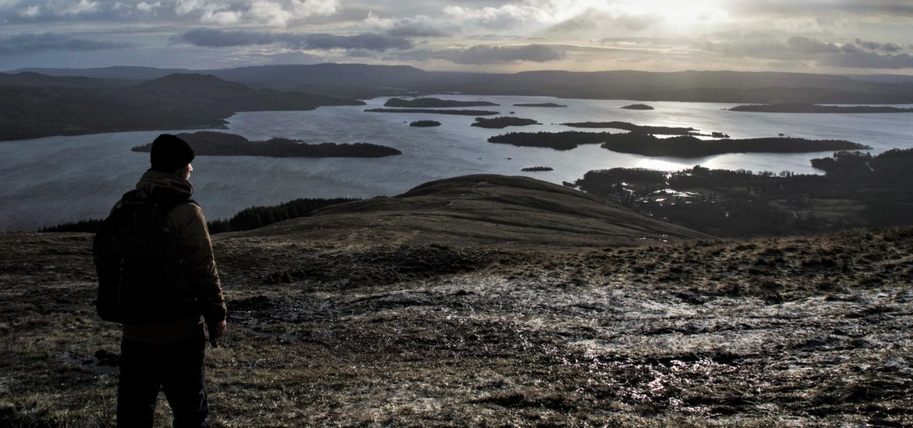 young-people-photo-competition-image-of-young-man-in-winter-clothes-on-hill-slope-admiring-panorama-of-loch-lomond-and-islands-dark-brown-and-gray-landscape-lit-up-by-winter-sun