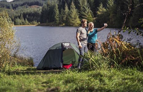 young-blond-couple-next-to-a-tent-taking-a-selfie-at-three-lochs-forest-drive-there-is-a-bike-in-the-foreground-and-loch-drunkie-and-forests-behind-them