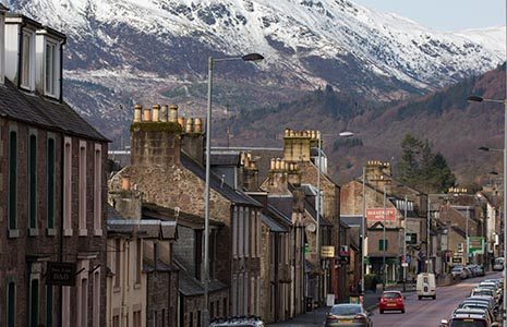 callander-main-street-with-old-buildings-facing-onto-street-cars-on-road-and-snowy-slopes-of-ben-ledi-mountain-in-the-background
