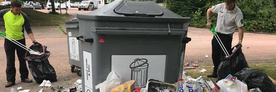 two-male-national-park-rangers-holding-bin-bags-and-picking-up-litter-using-sticks-next-to-overflowing-large-bins-in-luss