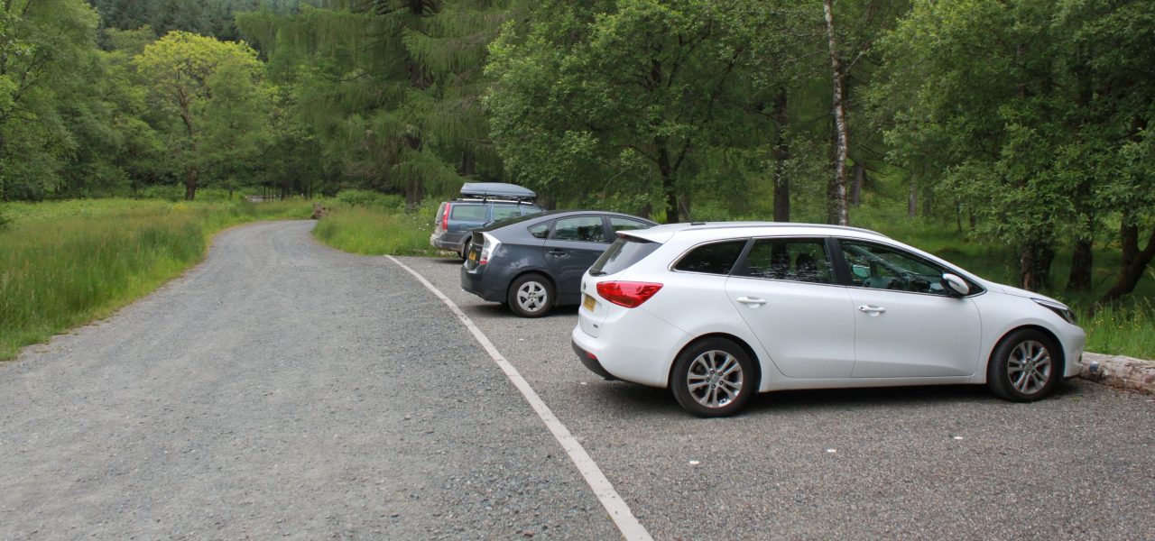 loch-chon-campsite-parking-spaces-next-to-level-track-with-three-cars-parked-on-the-right-of-the-picture-the-area-is-surrounded-by-lush-green-forest