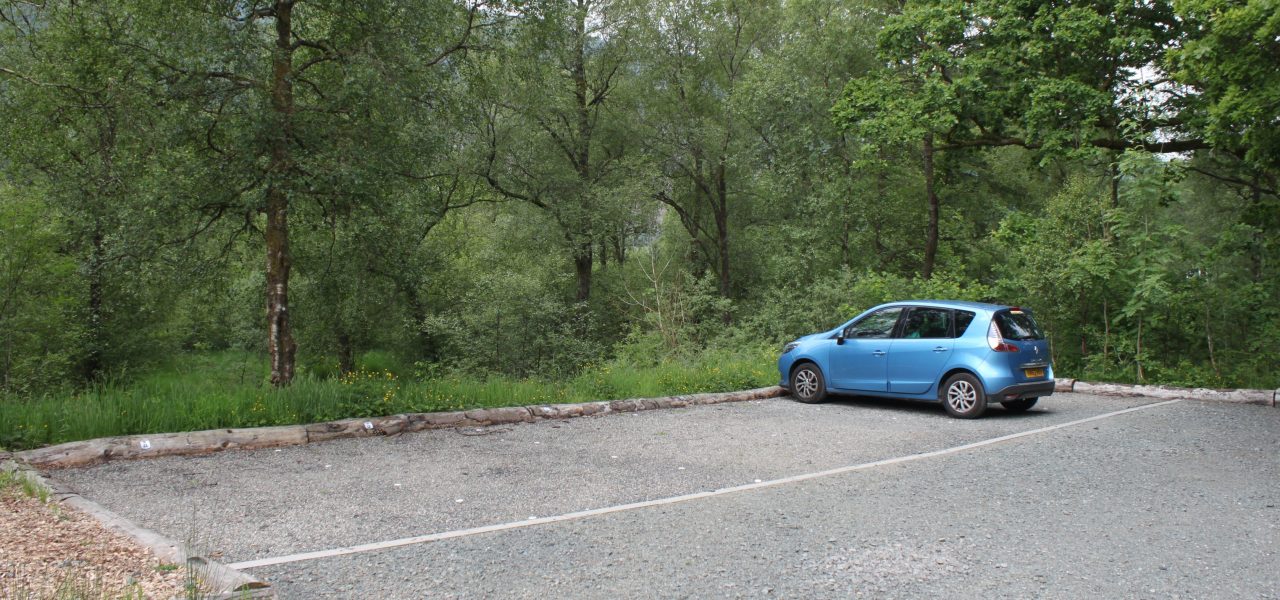 loch-chon-campsite-parking-spaces-next-to-level-track-with-one-blue-small-car-parked-on-the-right-of-the-picture-the-area-is-surrounded-by-lush-green-forest