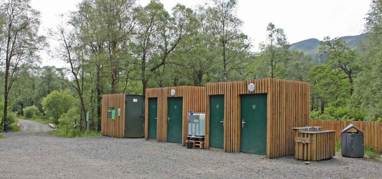 loch-chon-campsite-three-wooden-blocks-with-green-doors-the-two-on-the-right-for-toilets-a-water-tank-sink-and-bin-are-next-to-them-as-well-and-forest-of-the-campsite-behind-them