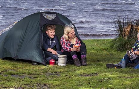 young-blond-couple-at-entrance-of-tent-next-to-gas-stove-and-pots-chatting-to-blonde-woman-holding-baby-at-entrance-of-another-tent-on-shores-of-loch-chon