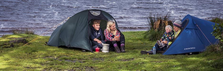 young-blond-couple-at-entrance-of-tent-next-to-gas-stove-and-pots-chatting-to-blonde-woman-holding-baby-at-entrance-of-another-tent-on-shores-of-loch-chon