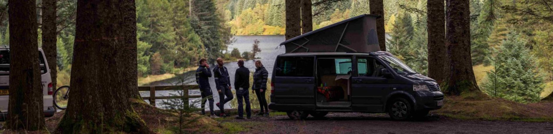 motorhome-campervan-national-park-three-man-and-a-woman-stand-outside-chatting-casually-stunning-view-of-loch-and-forests-behind-them
