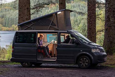 motorhome-parked-at-three-lochs-forest-drive-with-stunning-view-of-loch-and-coniferous-forests-around-the-door-is-open-and-two-men-sit-inside-one-is-reading-a-newspaper