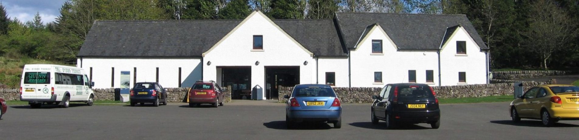 balmaha-visitor-centre-long-white-building-with-grey-slate-roof-and-with-trees-towering-behind-and-an-extensive-car-park-in-the-front