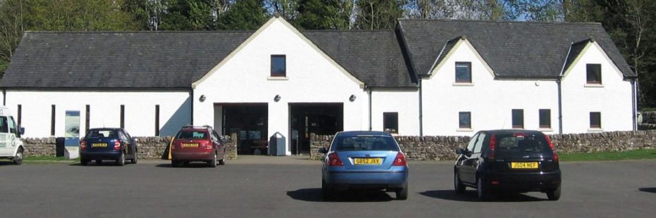 balmaha-visitor-centre-long-white-building-with-grey-slate-roof-and-with-trees-towering-behind-and-an-extensive-car-park-in-the-front